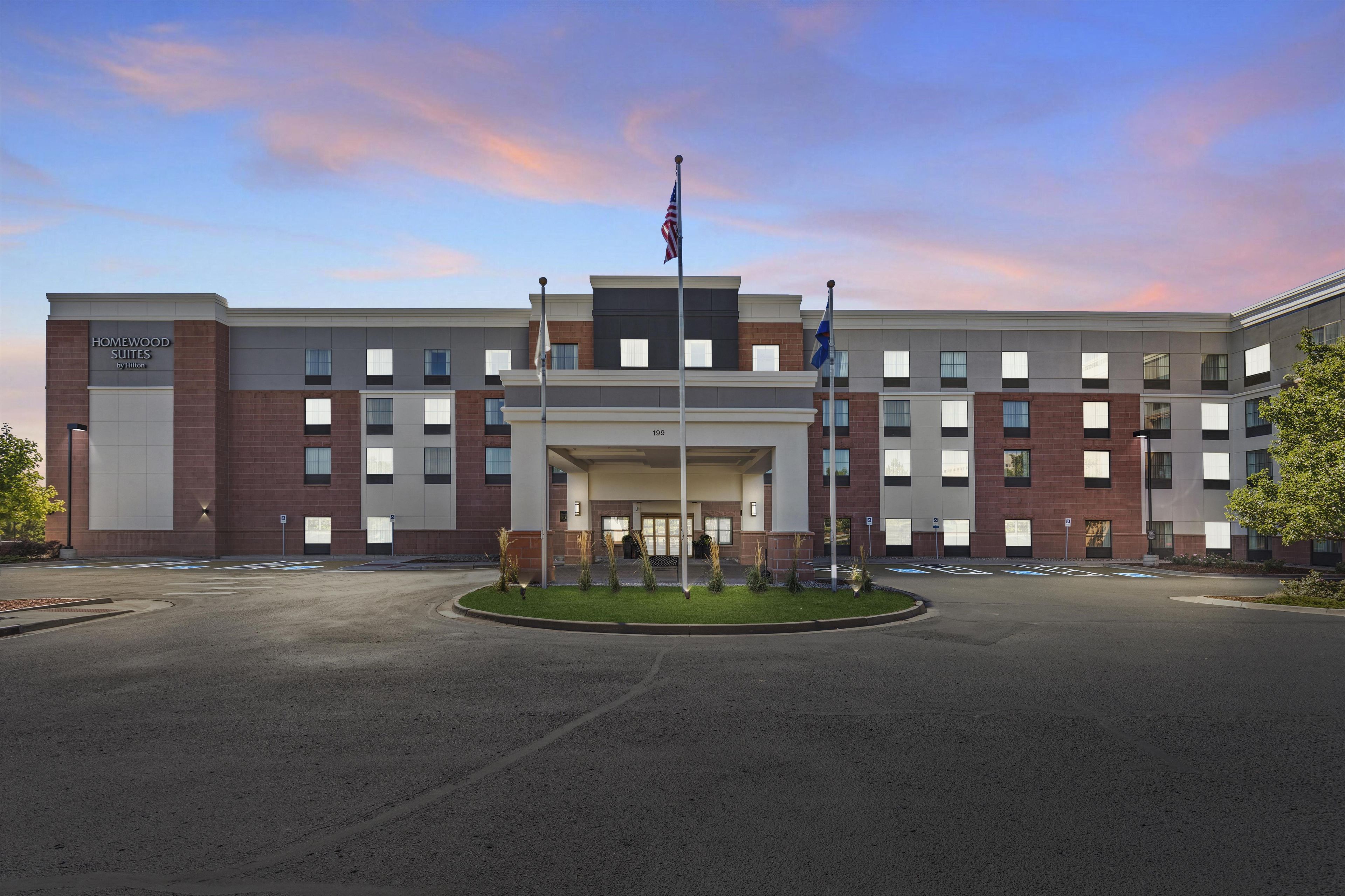A modern hotel building with a sign for 'Homewood Suites by Hilton' and a flagpole with flags.