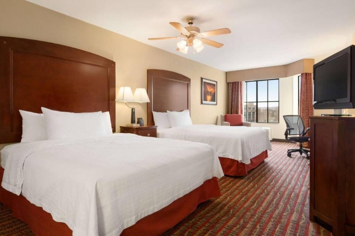 A hotel room with two beds, a ceiling fan, a TV, and a large window.
