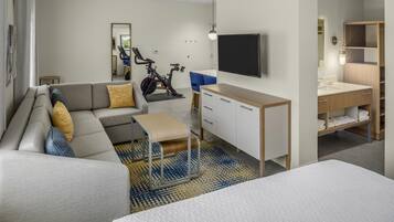 A modern hotel room with a sofa, a small table, a TV, a desk with drawers, and a bathroom with a sink and shelves.