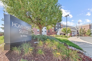 Señal de Homewood Suites by Hilton, rodeada por un jardín ajardinado con diversas plantas y un árbol grande.