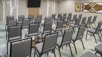 A conference room with rows of grey chairs, a large flat-screen TV on the wall, and two abstract paintings.