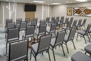 Un salón de conferencias con filas de sillas grises, un televisor de pantalla plana grande en la pared y dos cuadros abstractos.