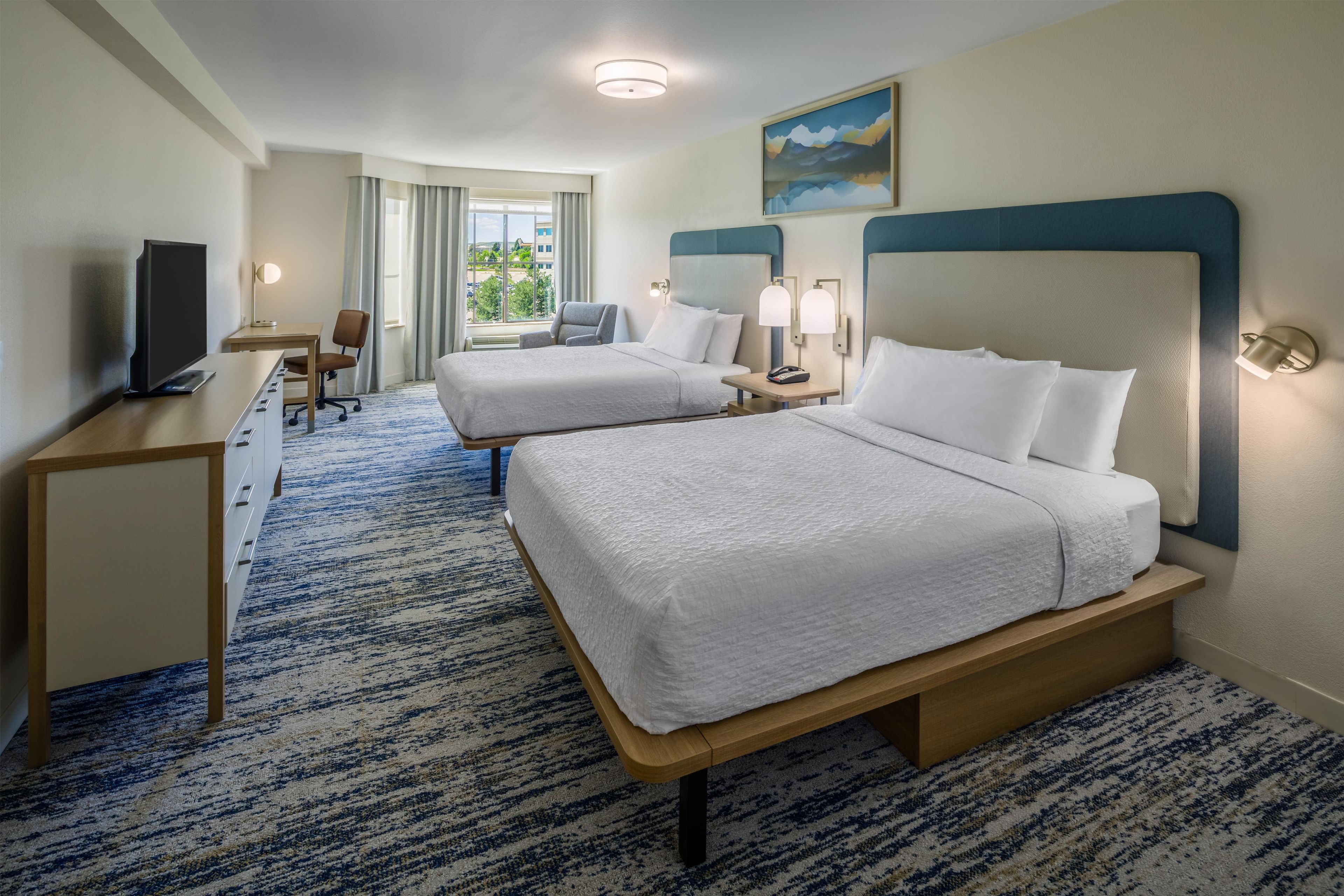 A hotel room with two beds, a desk, a television, and a window with curtains.