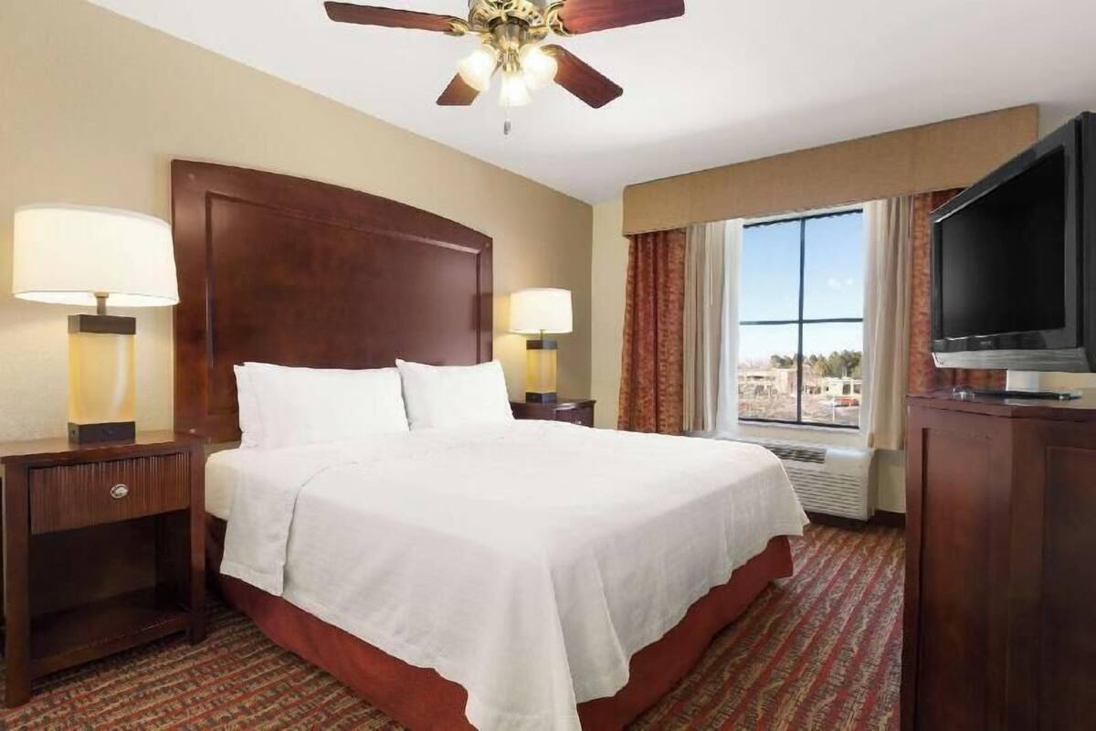 A hotel room with a large bed, two bedside lamps, a ceiling fan, a TV, and a window with a view.