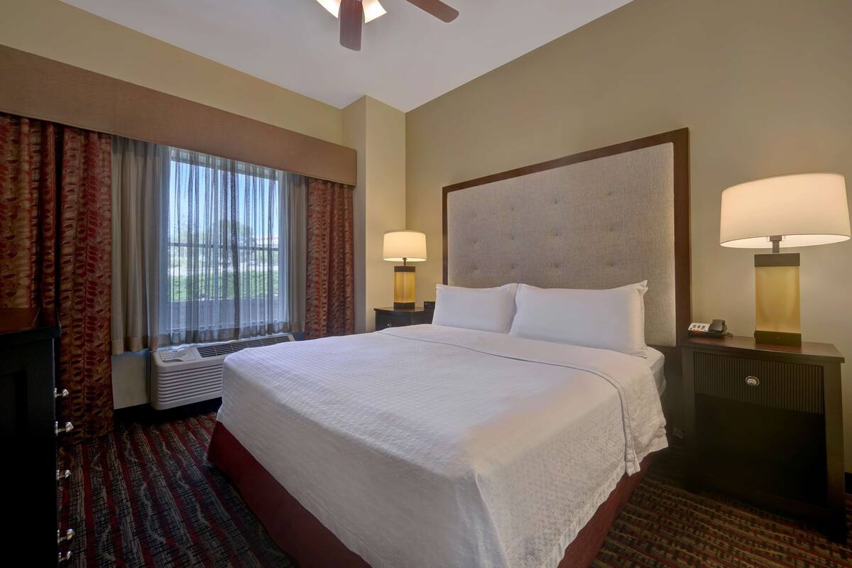 A hotel room with a large bed, two bedside lamps, a heater, and a window with curtains.