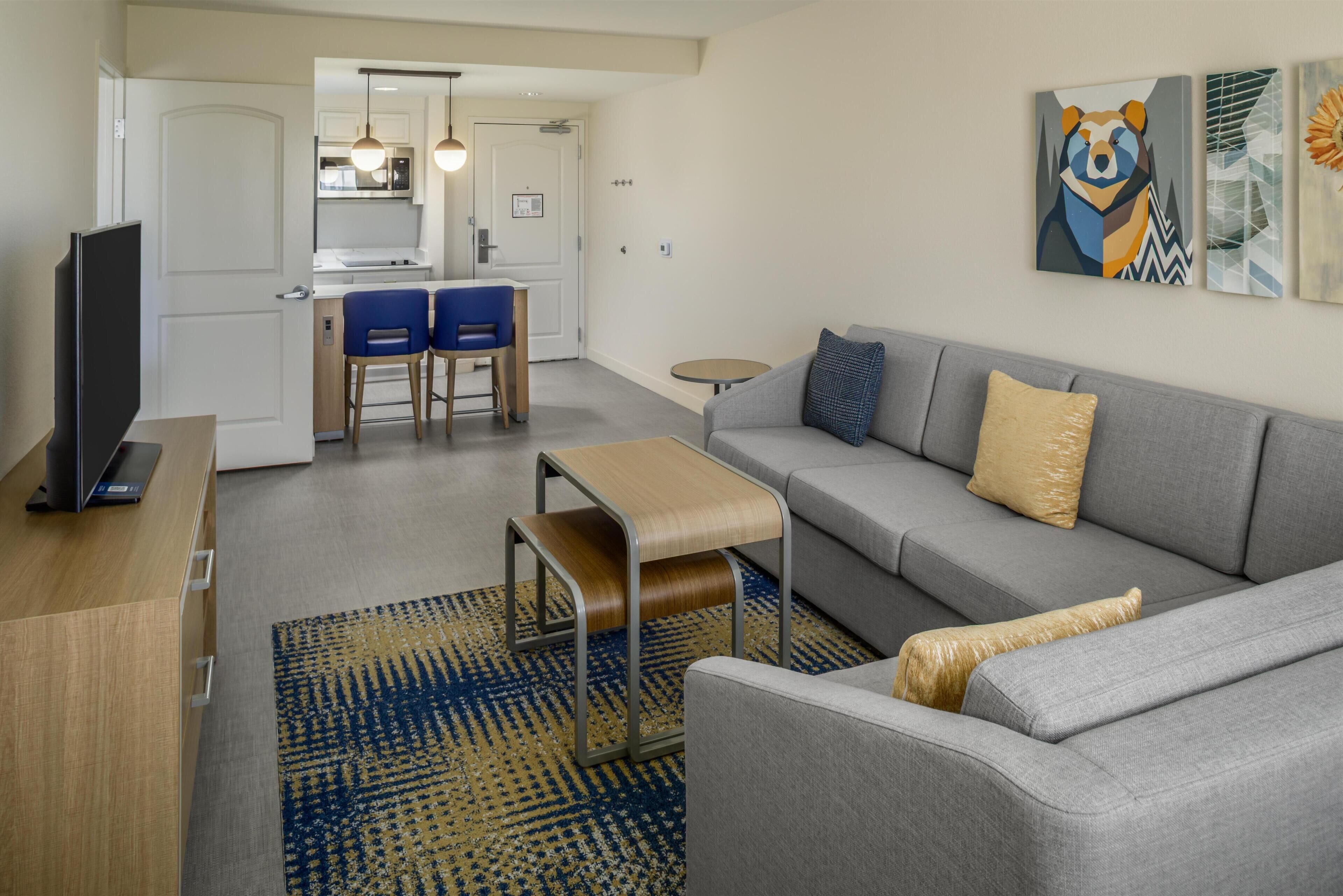 A modern living room with a grey sofa, a small wooden coffee table, and a TV on a wooden cabinet. There is a kitchen in the background with a dining area featuring two blue chairs.