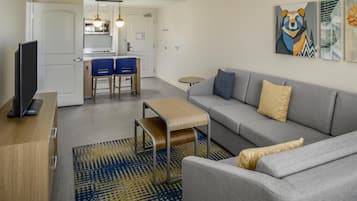 A modern living room with a grey sofa, a small wooden coffee table, and a TV on a wooden cabinet. There is a kitchen in the background with a dining area featuring two blue chairs.