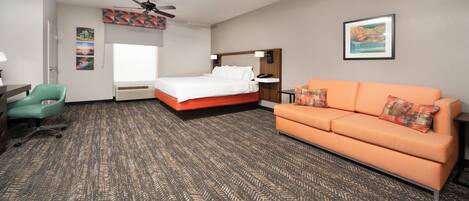 A hotel room with a bed, an orange sofa, a teal chair, a desk with a lamp, and a ceiling fan.