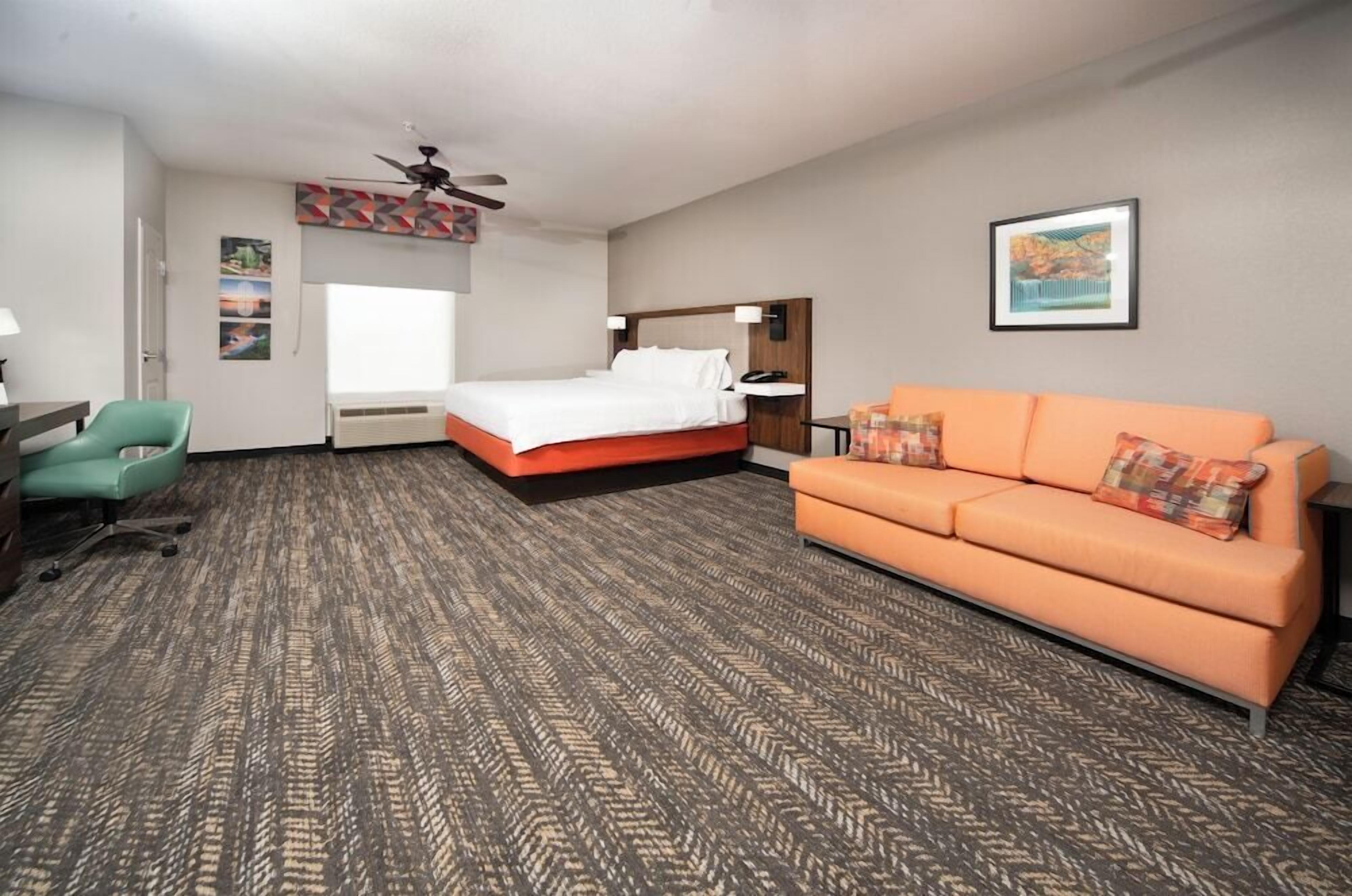 Habitación de hotel con una cama, sofá naranja, escritorio con silla, ventilador de techo y obras de arte en las paredes.