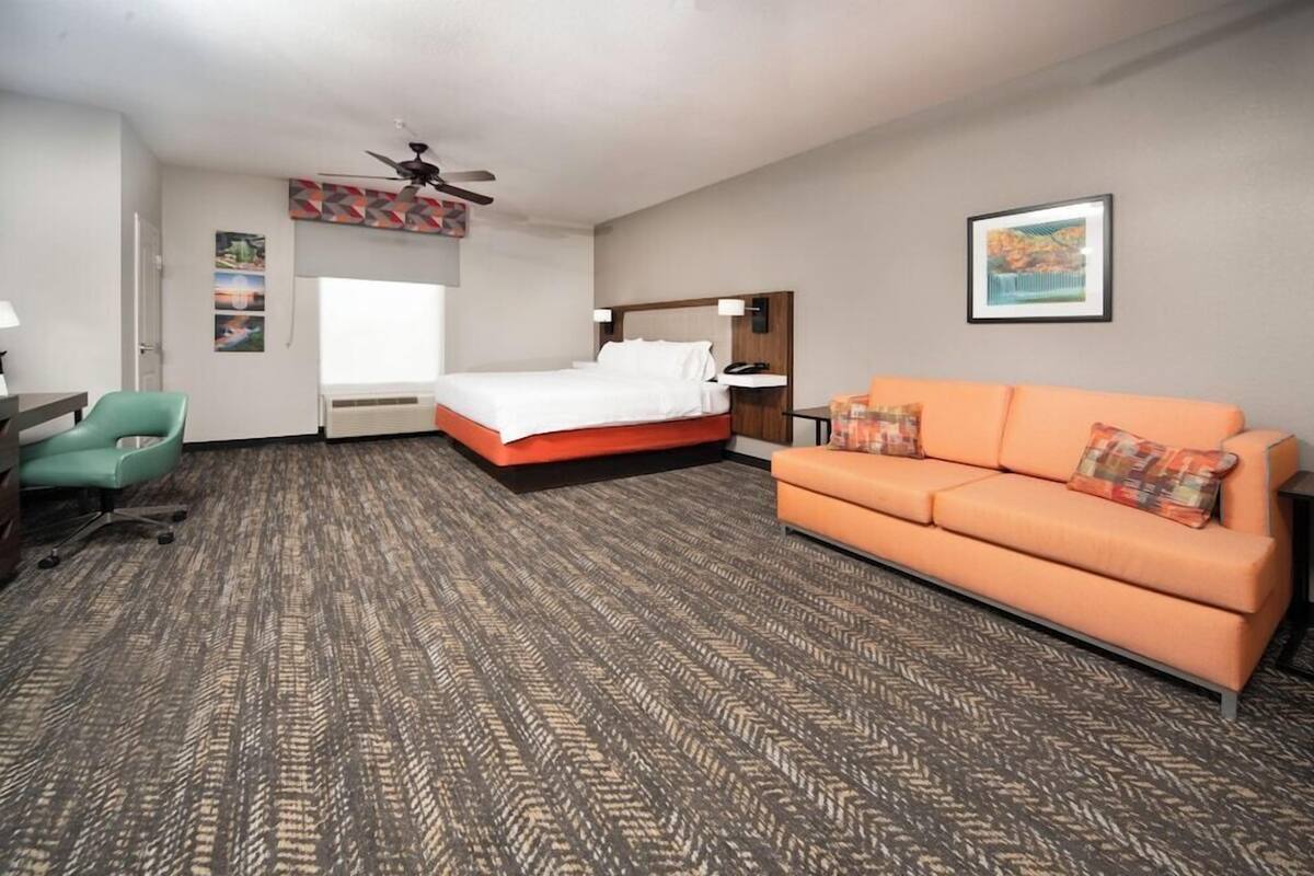 A hotel room with a bed, an orange sofa, a desk with a chair, a ceiling fan, and artwork on the walls.