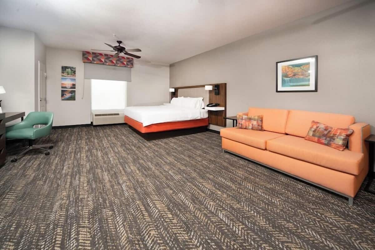 Habitación de hotel con una cama, sofá naranja, escritorio con silla, ventilador de techo y obras de arte en las paredes.