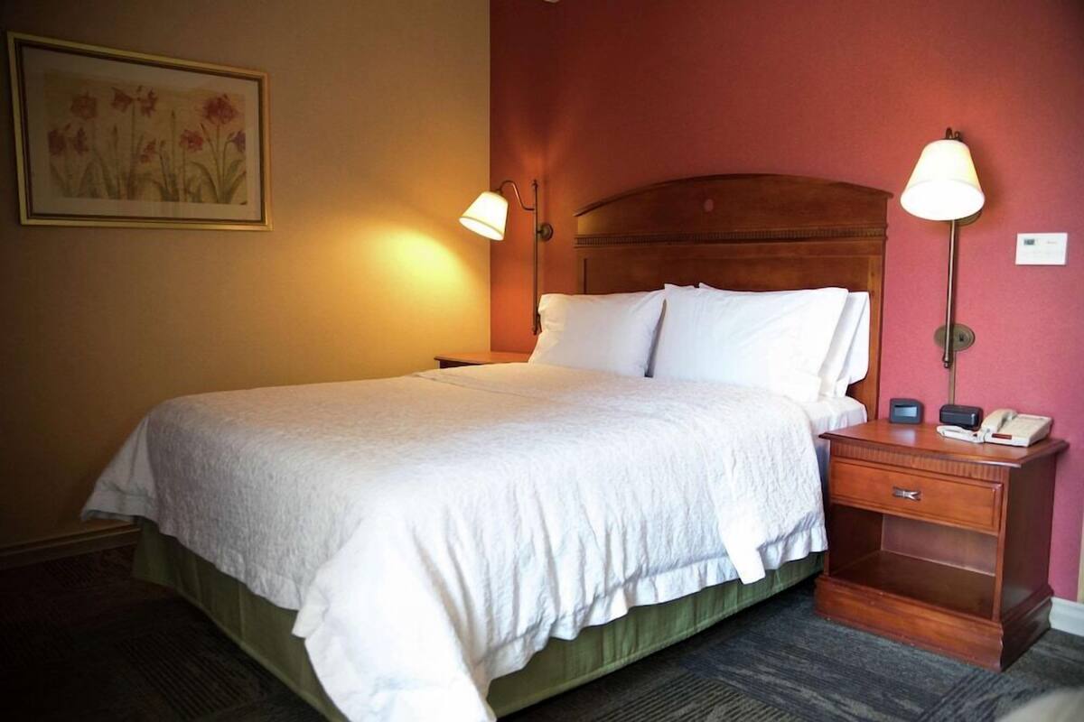 A neatly made bed with white linens, a wooden headboard, two bedside lamps, a nightstand with a phone and a small table, and a framed floral artwork on the wall.