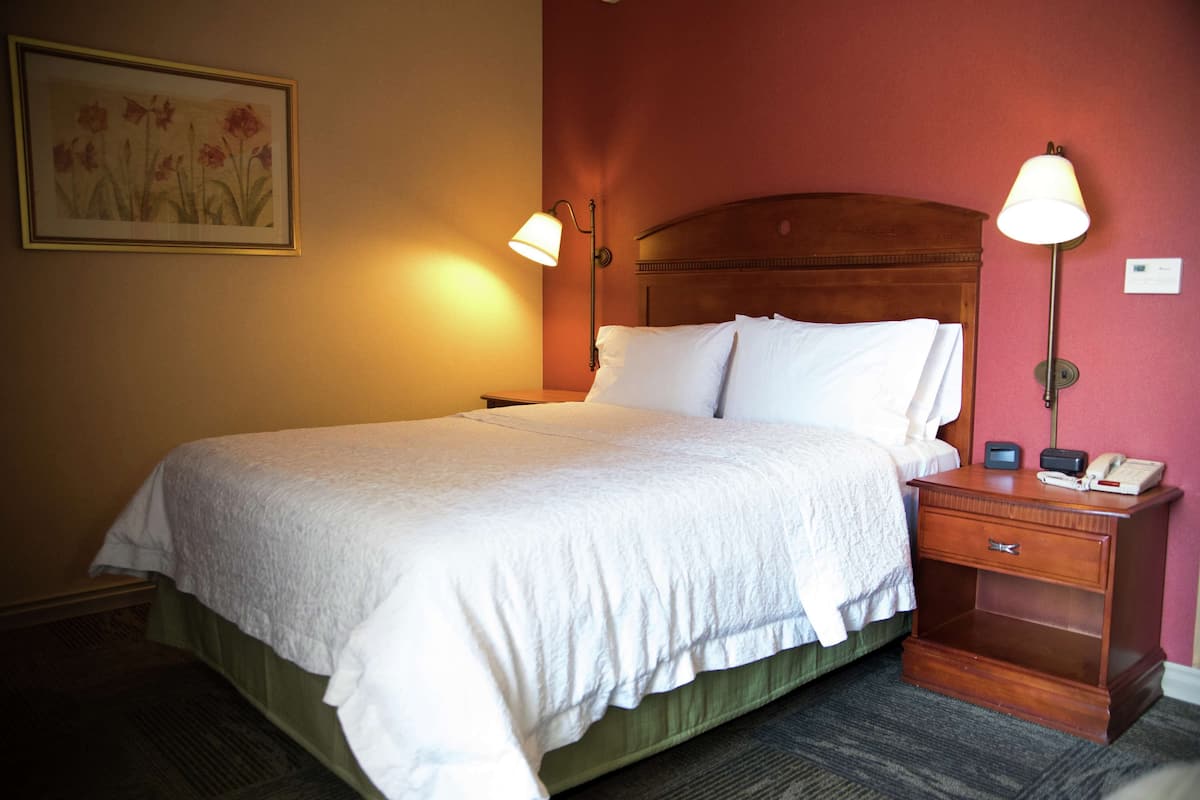 A neatly made bed with white linens, two bedside lamps, a nightstand with a phone and a small clock, and a framed floral artwork on the wall.