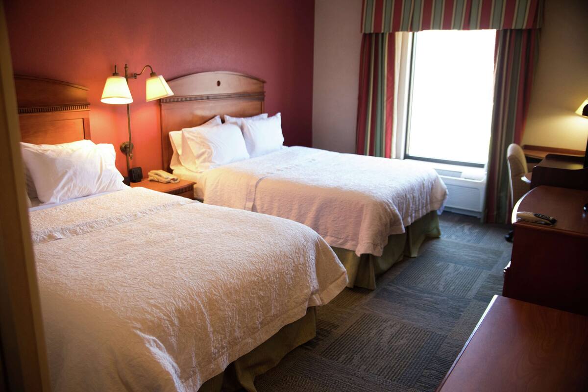 A hotel room with two beds, a desk, a chair, and a window with curtains.