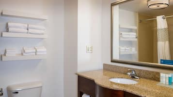 A hotel bathroom with a toilet, sink, and mirror. There are shelves with folded towels above the toilet and a towel rack in the shower area.