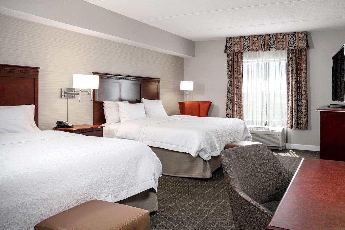 A hotel room with two beds, a desk, a chair, a TV, and a window with curtains.