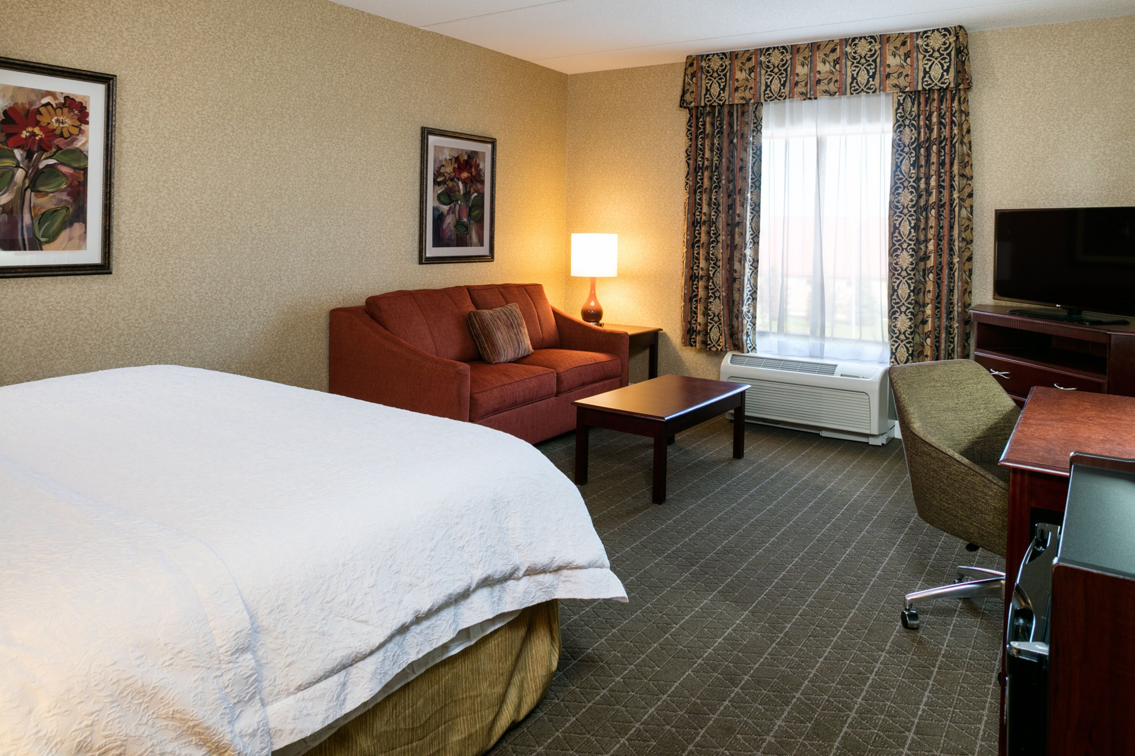 A hotel room with a large bed, a sofa, a small table, a TV, and a window with curtains.