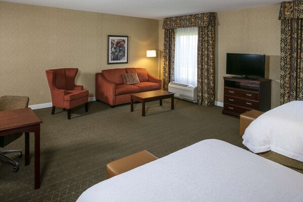 Una habitación de hotel con cama, sofá, sillón, escritorio, televisor y ventana con cortinas.