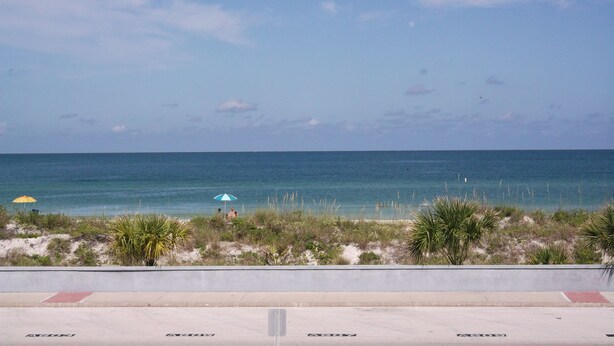 A beach view with a clear sky, a sandy area with vegetation, and a few umbrellas. - Camelot by the Sea by Liberte' (St. Pete Beach)