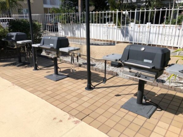 An outdoor barbecue area with grills and tables, surrounded by a white fence and palm trees.
