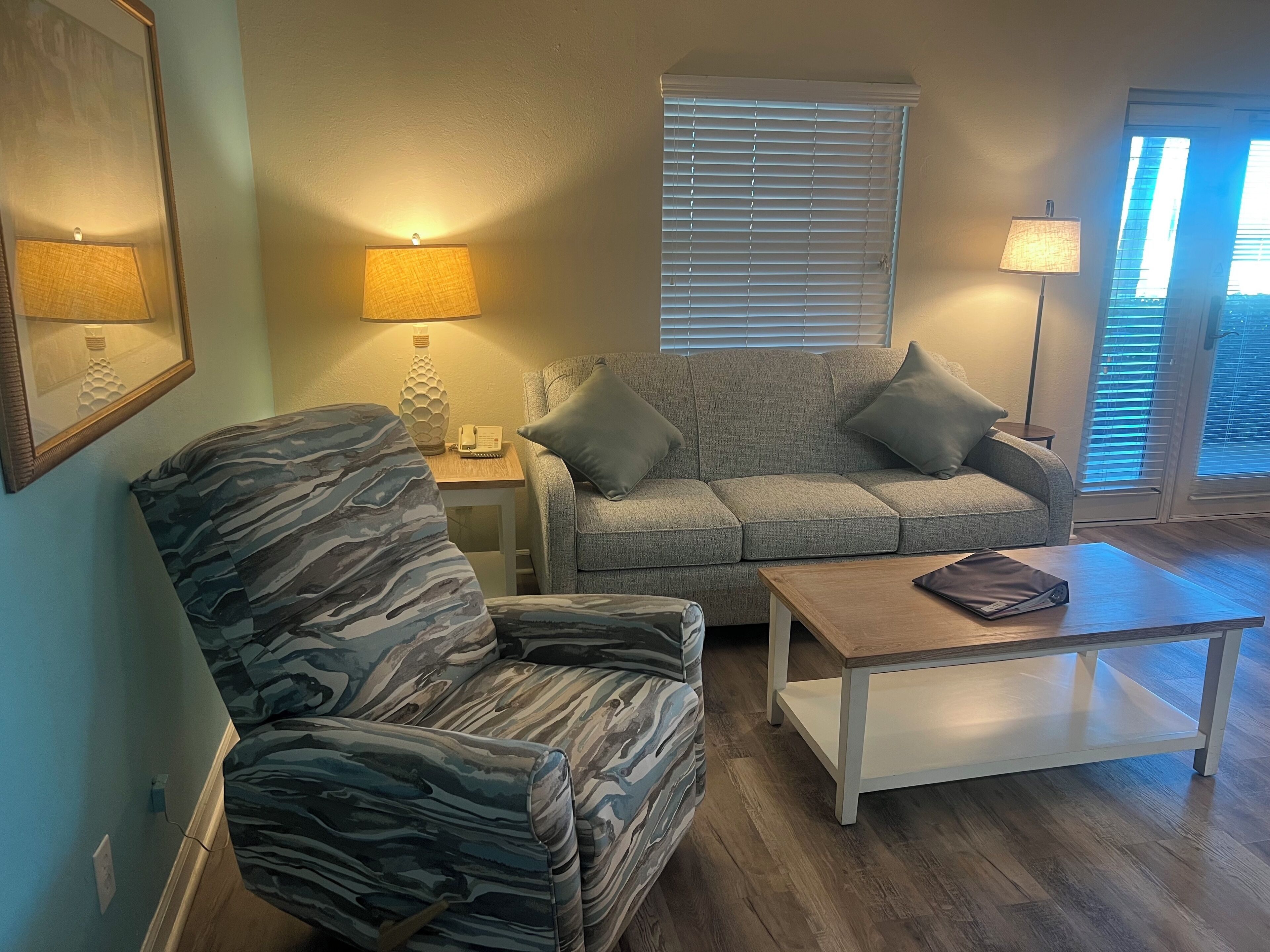 A living room with a patterned armchair, a grey sofa, a wooden coffee table, and a lamp on a side table.