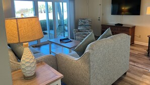 A living room with a sofa, a table, a lamp, a television, and a sliding glass door leading to an outdoor area with chairs. - Camelot by the Sea by Liberte' (St. Pete Beach)