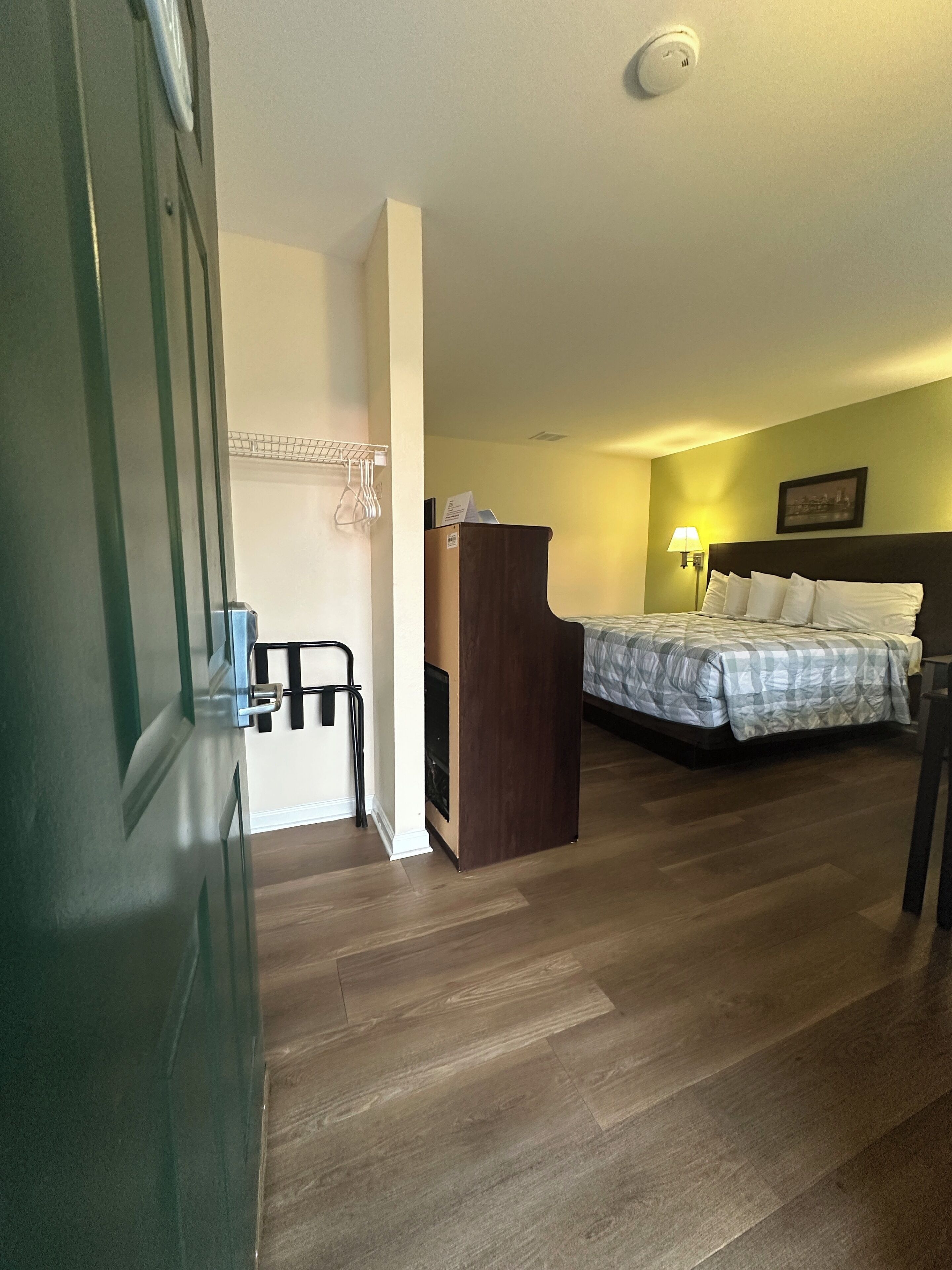 Een hotelkamer met een houten vloer, een bed met een geruit sprei, een bureau met stoel, een wandplank en een wandlamp.