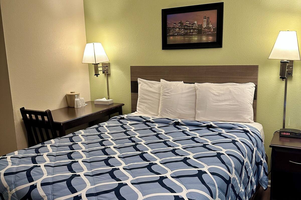 A hotel room with a bed, two bedside lamps, a nightstand, a chair, and a framed picture on the wall.