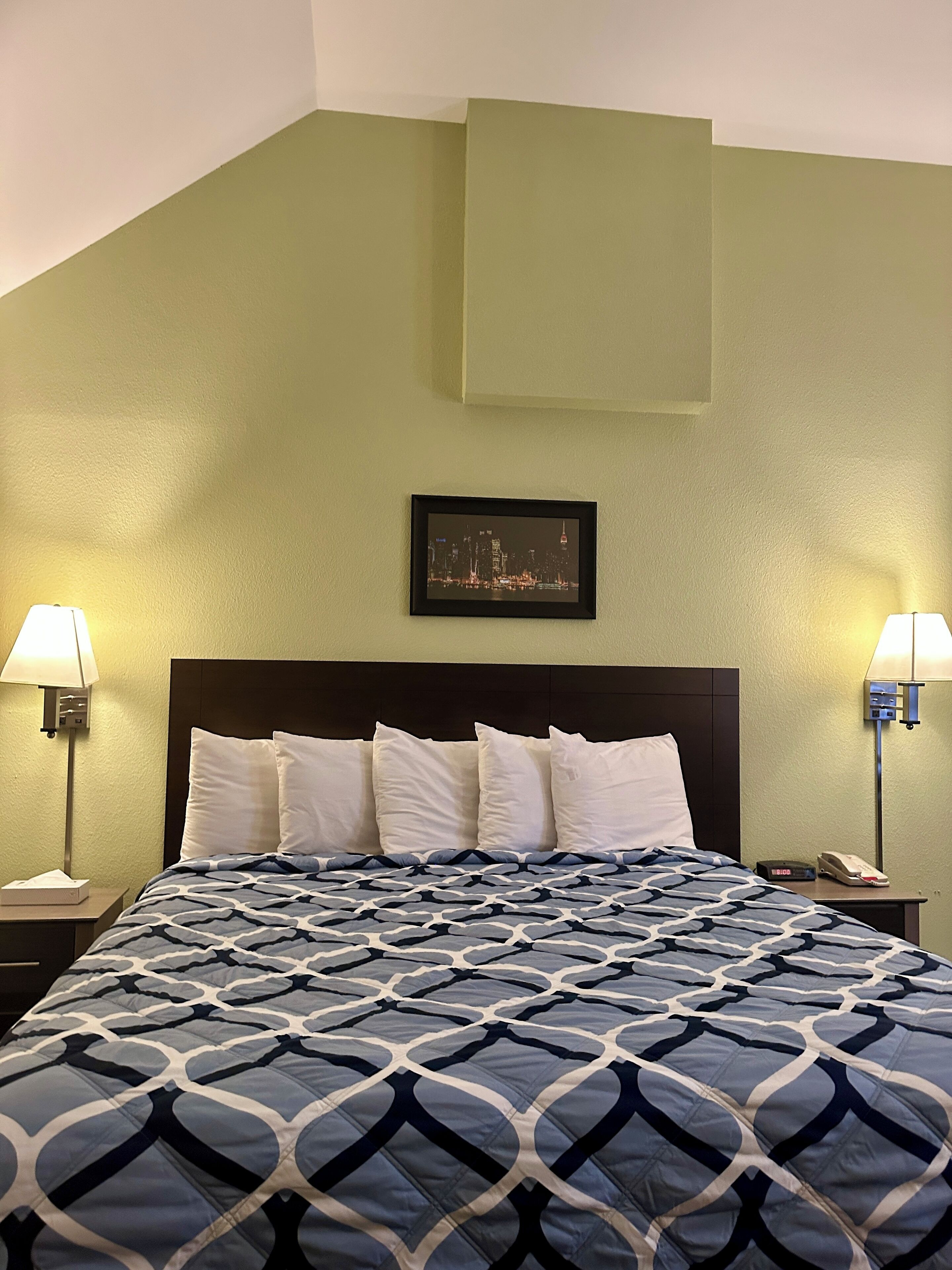 A hotel room with a bed, two bedside tables, a nightstand, a framed picture on the wall, and a clock.