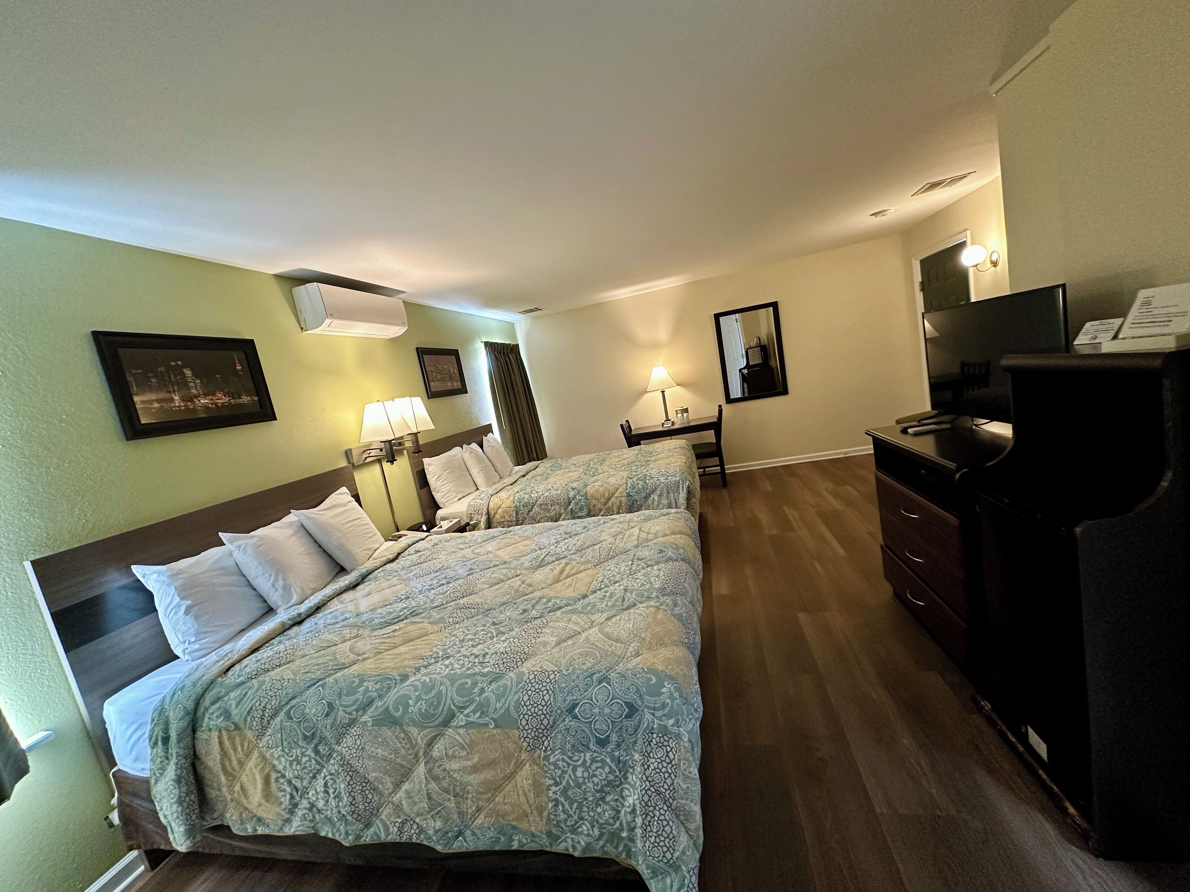A hotel room with two beds, a desk, a dresser, a TV, and a framed picture on the wall.