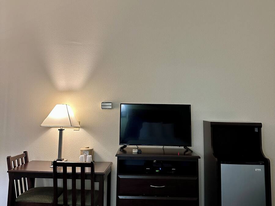 A small room with a wooden table, a lamp, a flat-screen TV, a dark-colored entertainment unit, and a refrigerator.