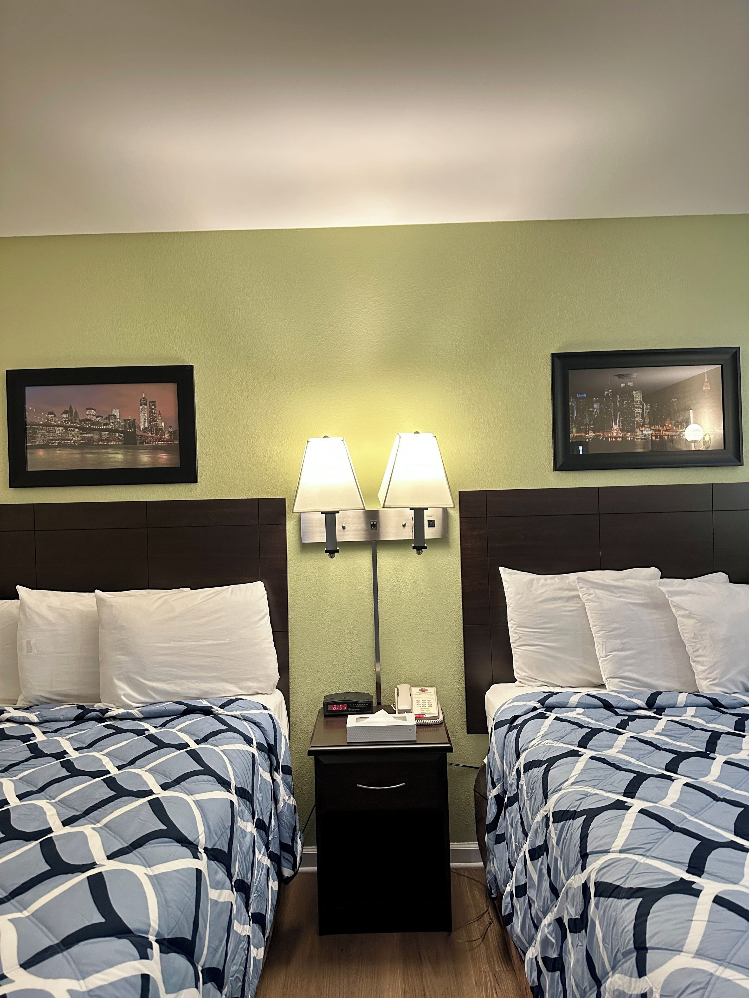 A hotel room with two beds, a nightstand with a lamp, a clock, and a phone.