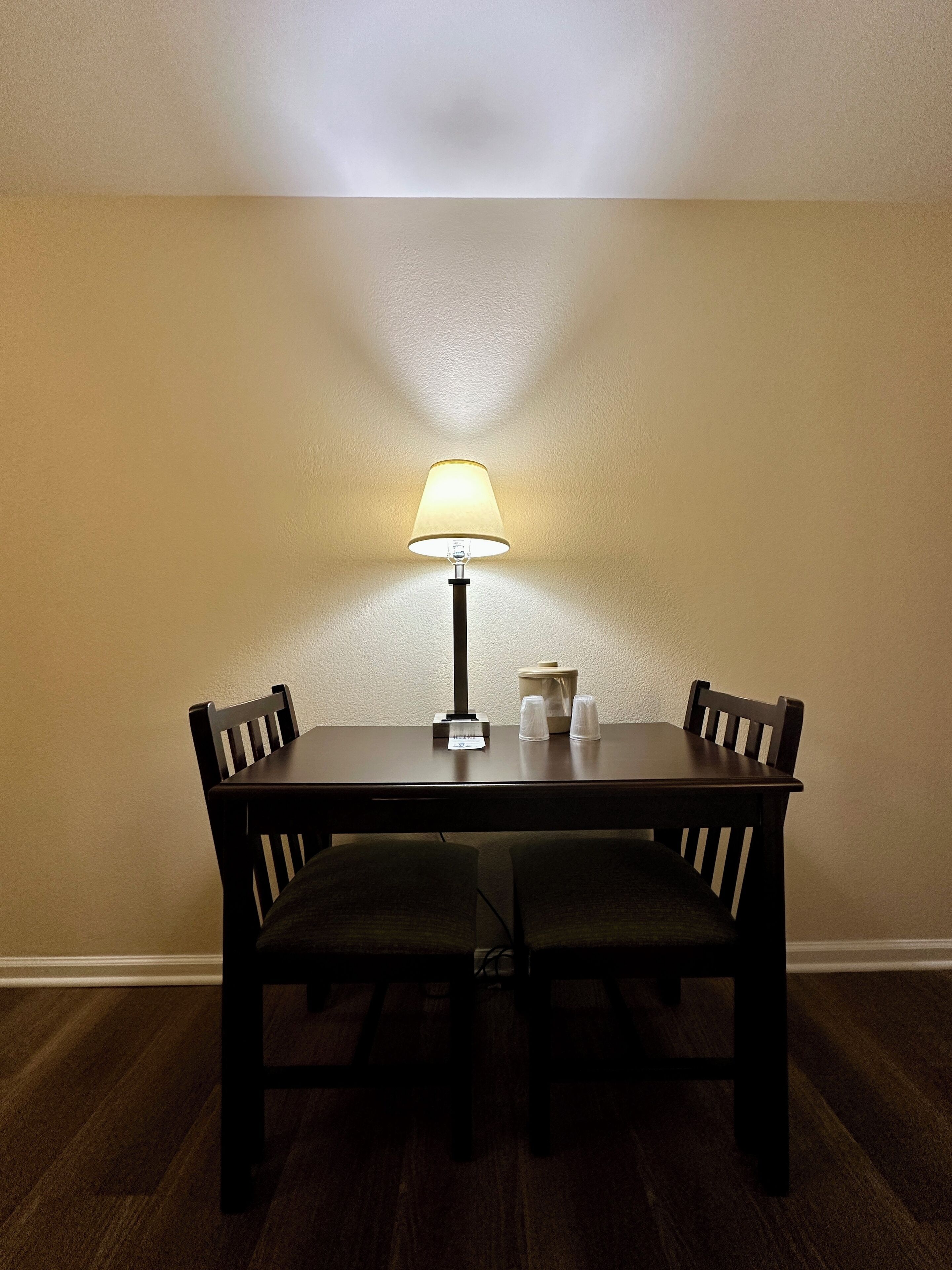 Ett matbord med två stolar, en lampa och ett litet bord med koppar och en näsdukslåda.