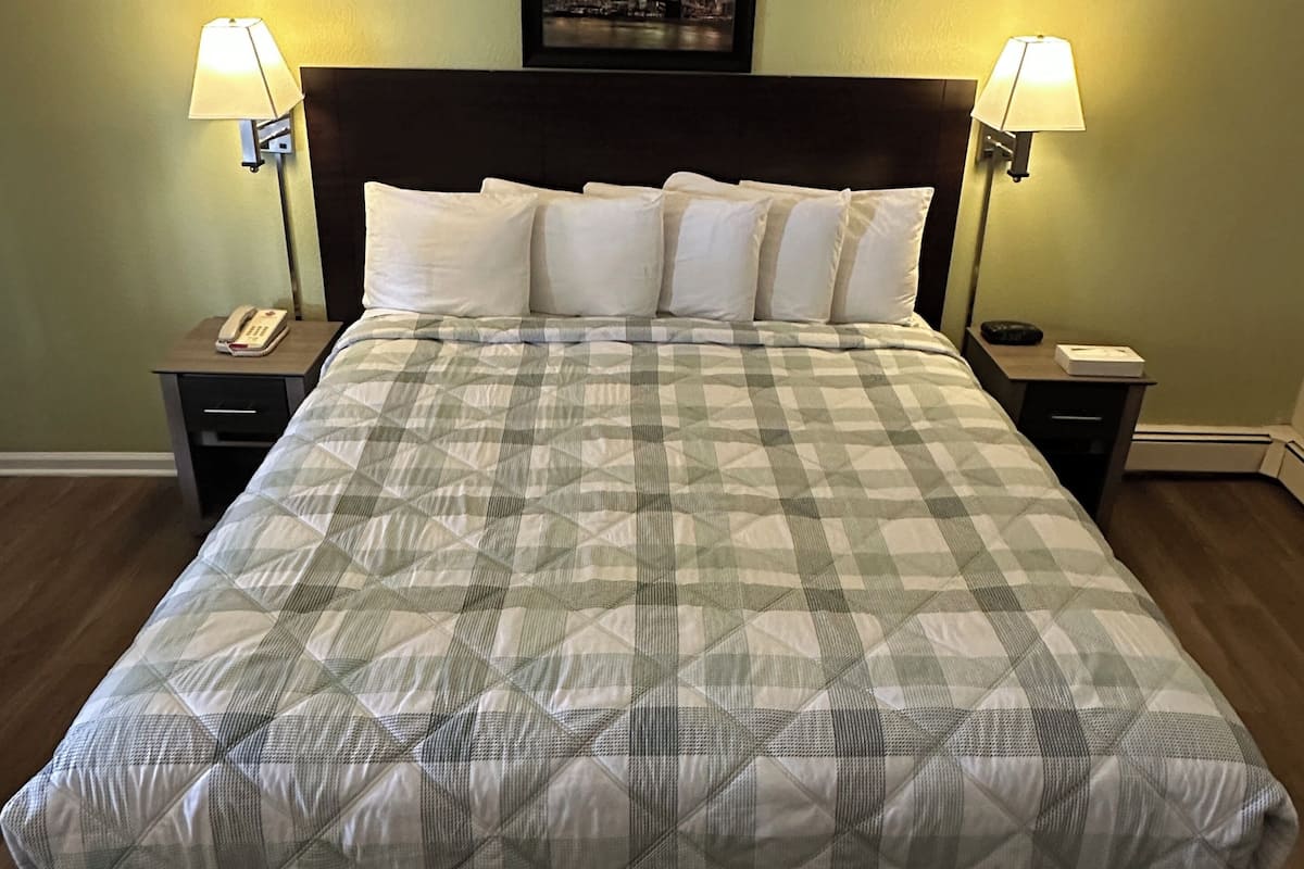 A neatly made bed with a checkered bedspread, two bedside lamps, a framed picture above the headboard, and a nightstand with a phone and a remote control.