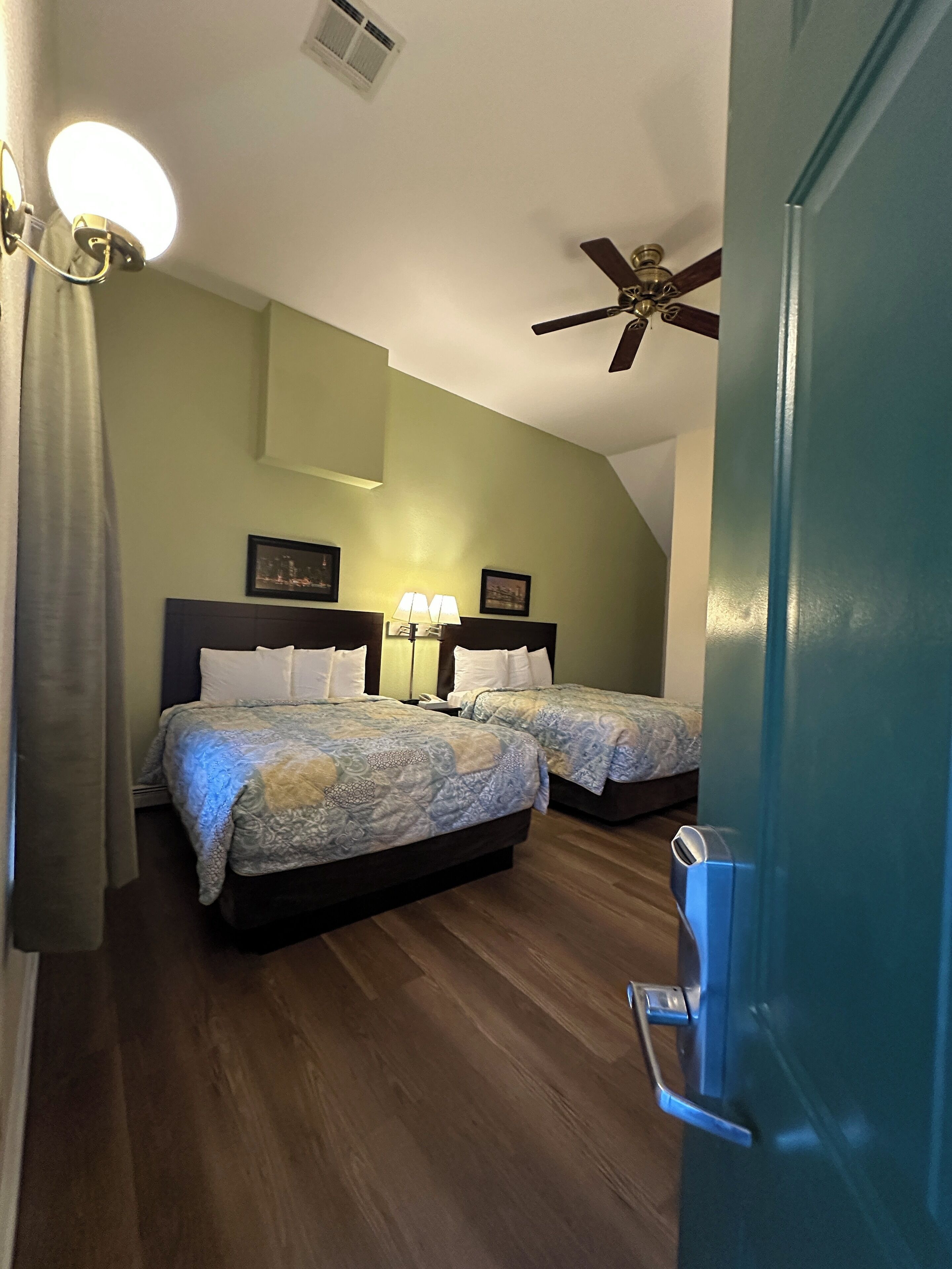 Quarto de hotel com duas camas, um ventilador de teto, uma mesinha de cabeceira com abajur e uma porta com puxador.