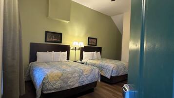 Quarto de hotel com duas camas, um ventilador de teto, uma mesinha de cabeceira com abajur e uma porta com puxador.