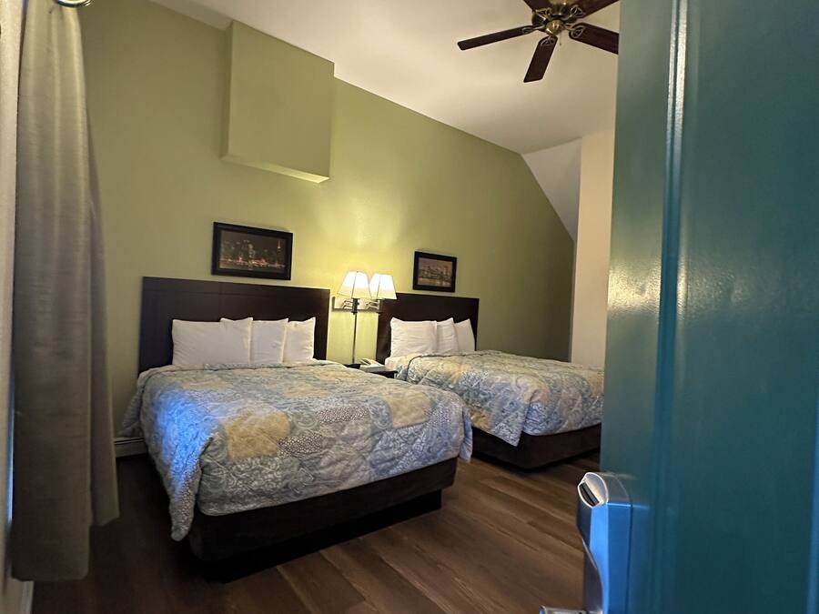 Quarto de hotel com duas camas, um ventilador de teto, uma mesinha de cabeceira com abajur e uma porta com puxador.