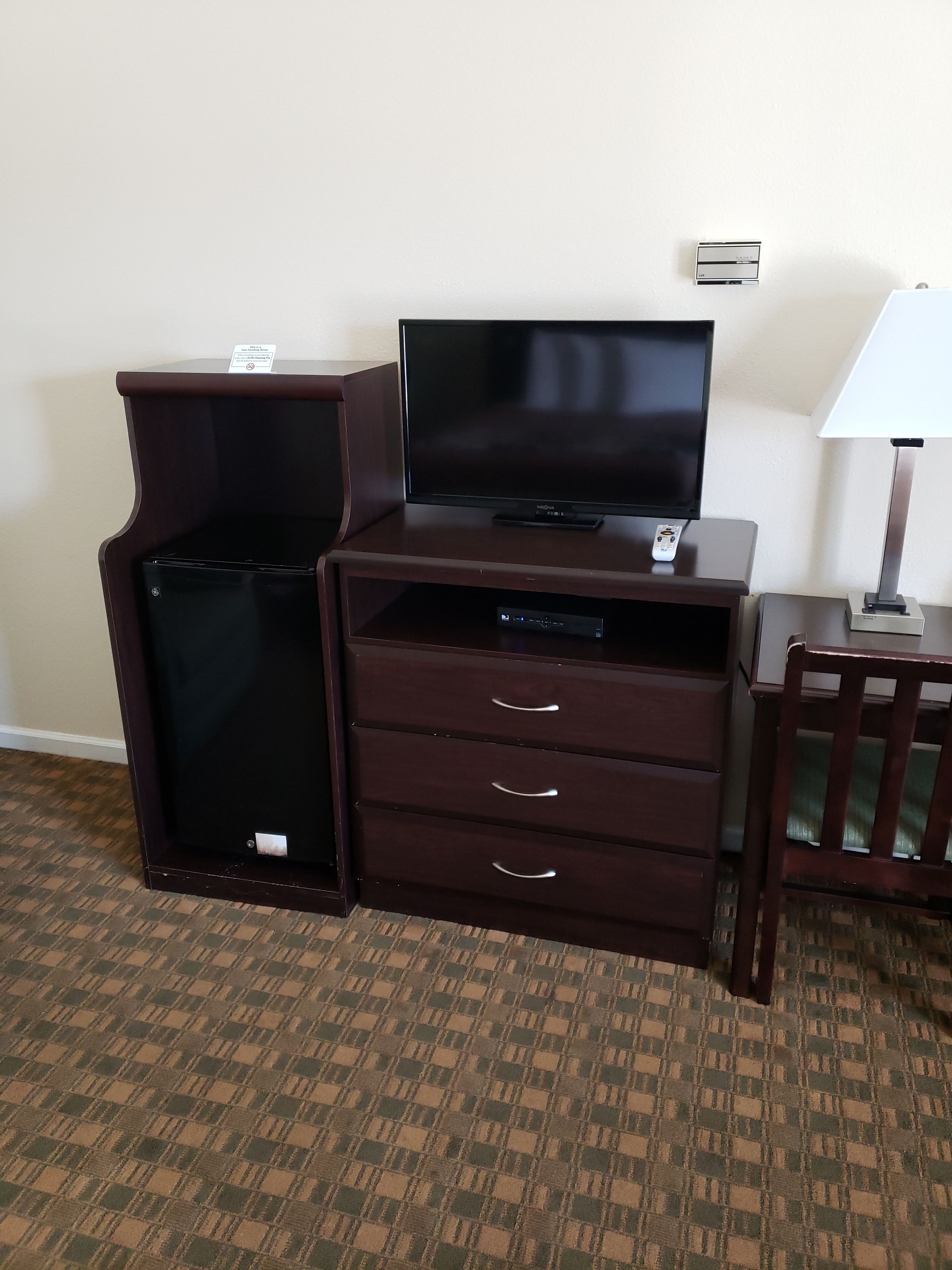 A compact hotel room with a flat-screen TV on a wooden TV stand, a small desk with a lamp, and a chair.