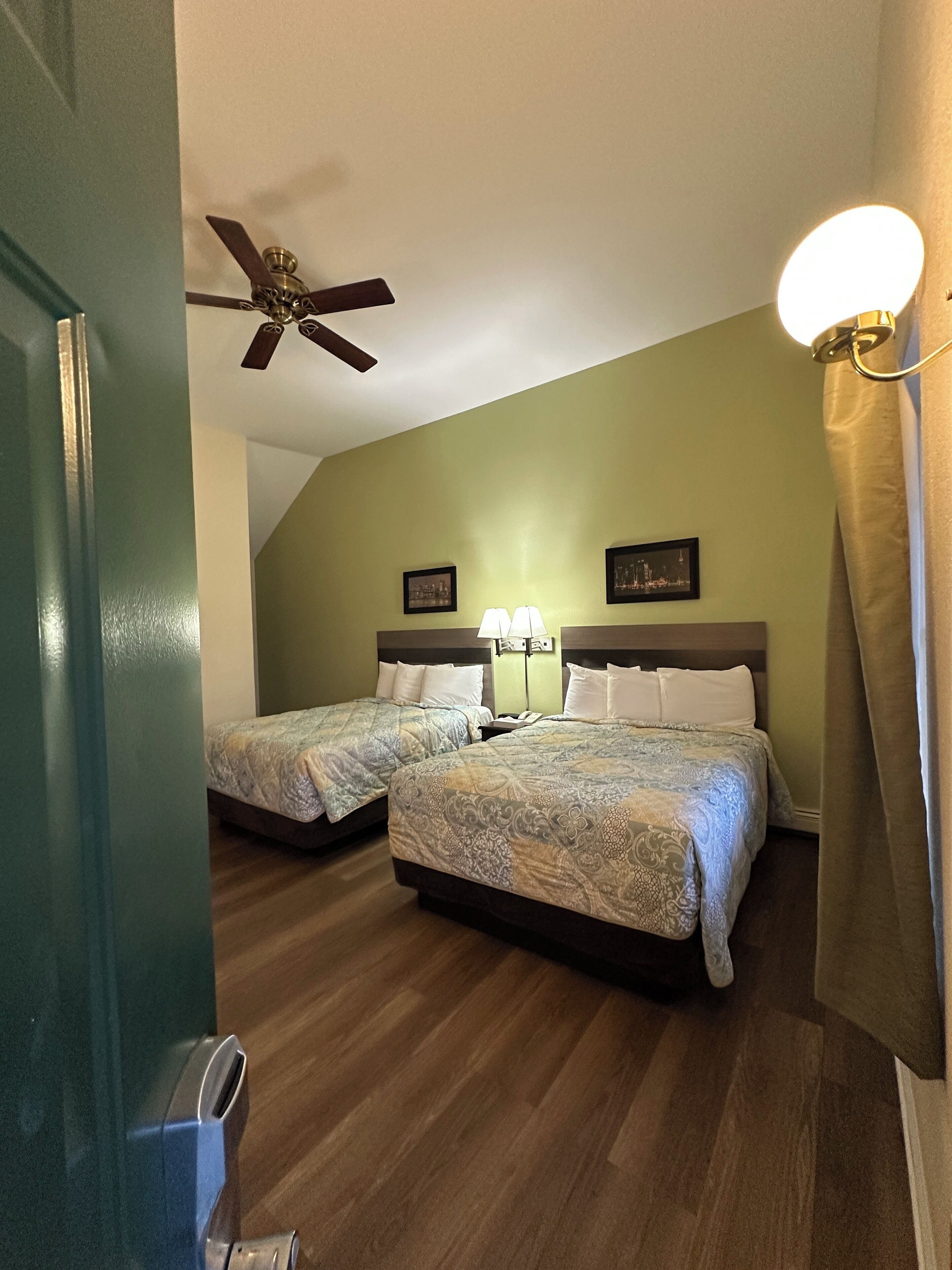 A hotel room with two beds, a ceiling fan, a nightstand with a lamp, and a picture on the wall.