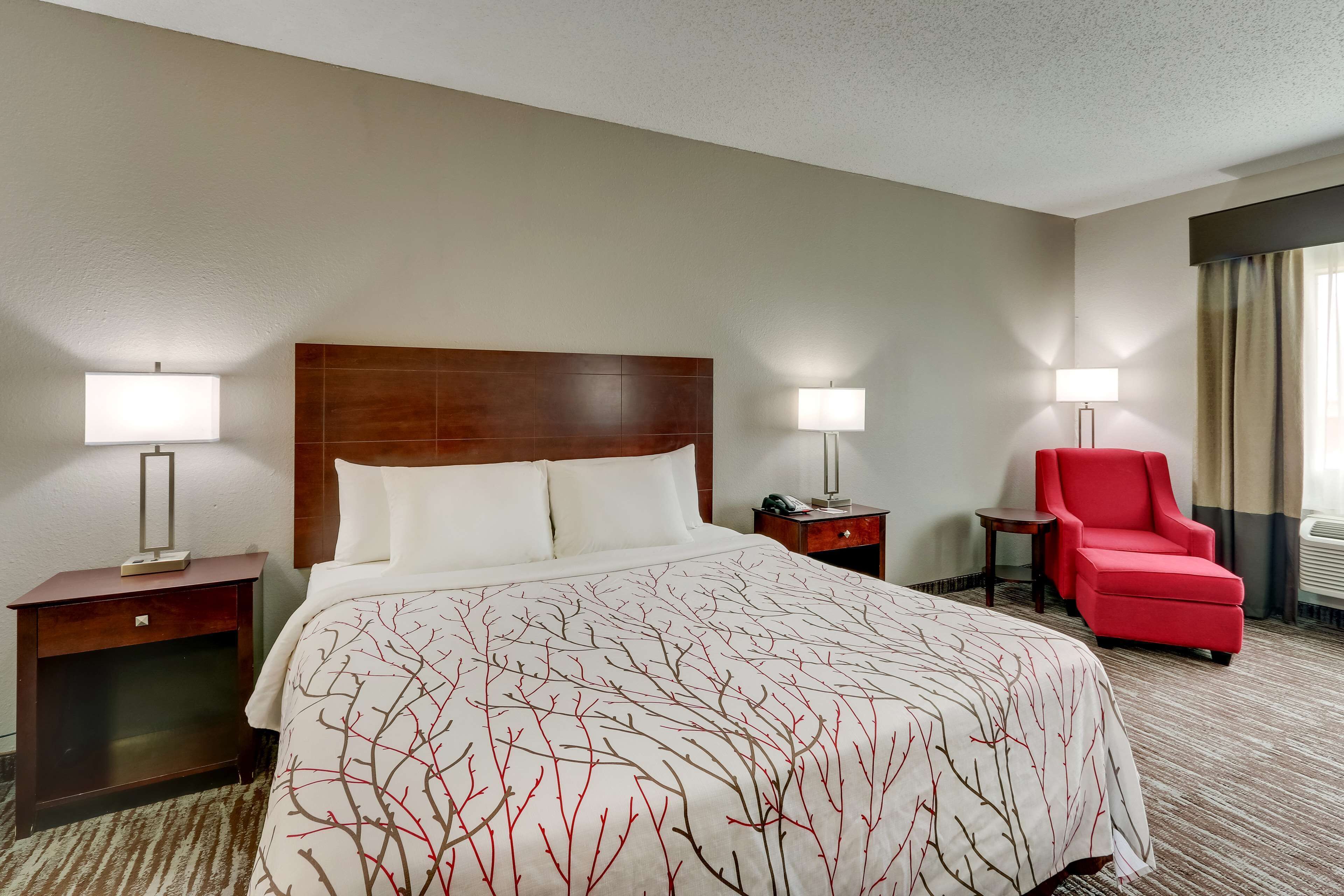 Quarto de hotel com uma cama grande, duas mesas de cabeceira com luminárias, uma poltrona vermelha e uma janela com cortinas.