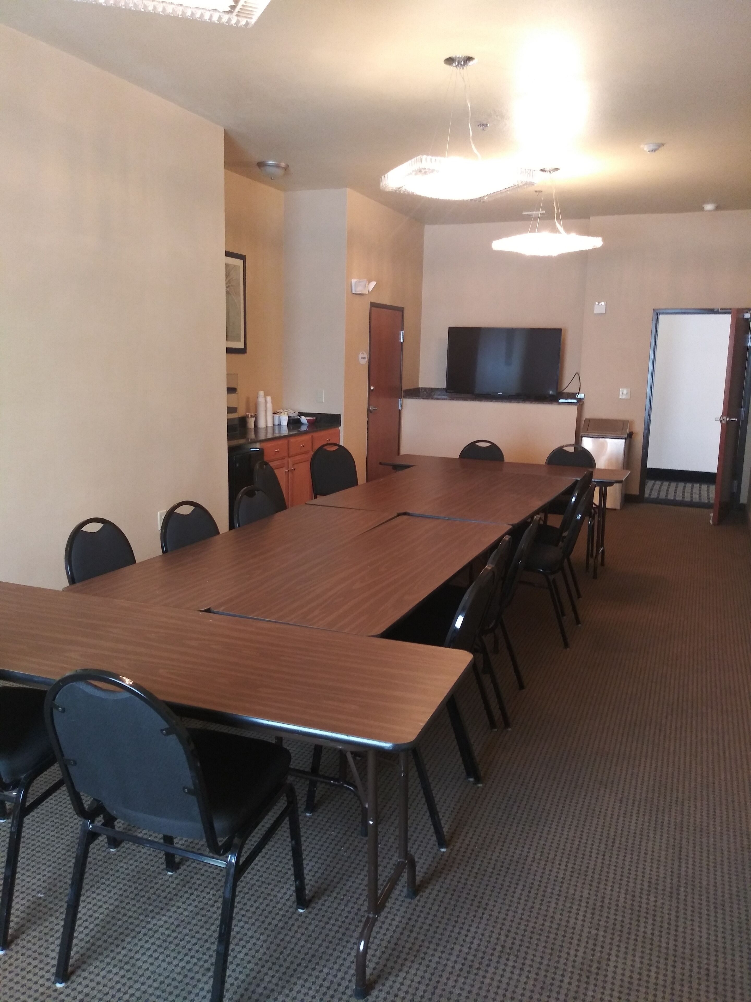 A conference room with a long wooden table, black chairs, a flat-screen TV mounted on the wall, and a kitchenette area with cabinets and a countertop.