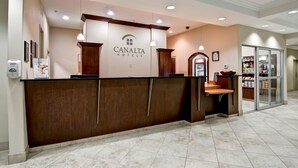 Una mesa de recepción en CANALTA HOTELS con un mostrador de madera, una pantalla digital y una puerta de cristal que da a una zona de máquinas expendedoras.