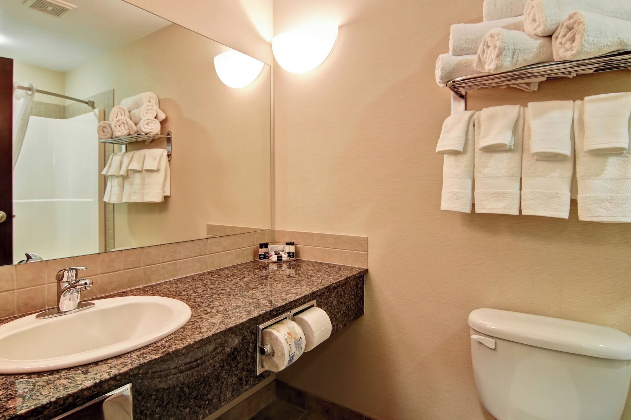 A bathroom with a granite countertop sink, a mirror, a toilet, and towels neatly arranged on a rack.