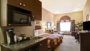 Habitación de hotel con kitchenette, una cama con colcha, escritorio, silla, televisor y ventana con cortinas.