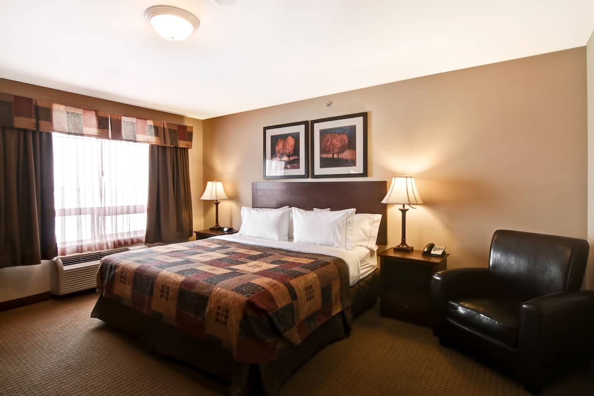 A hotel room with a large bed, two bedside lamps, a chair, a nightstand with a phone, and a window with curtains.