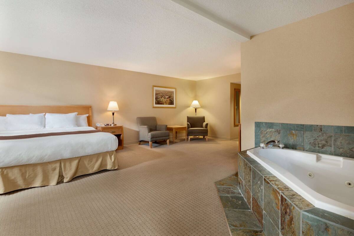 Habitación de hotel con una cama grande, dos sillas, una mesita, una lámpara y una bañera.