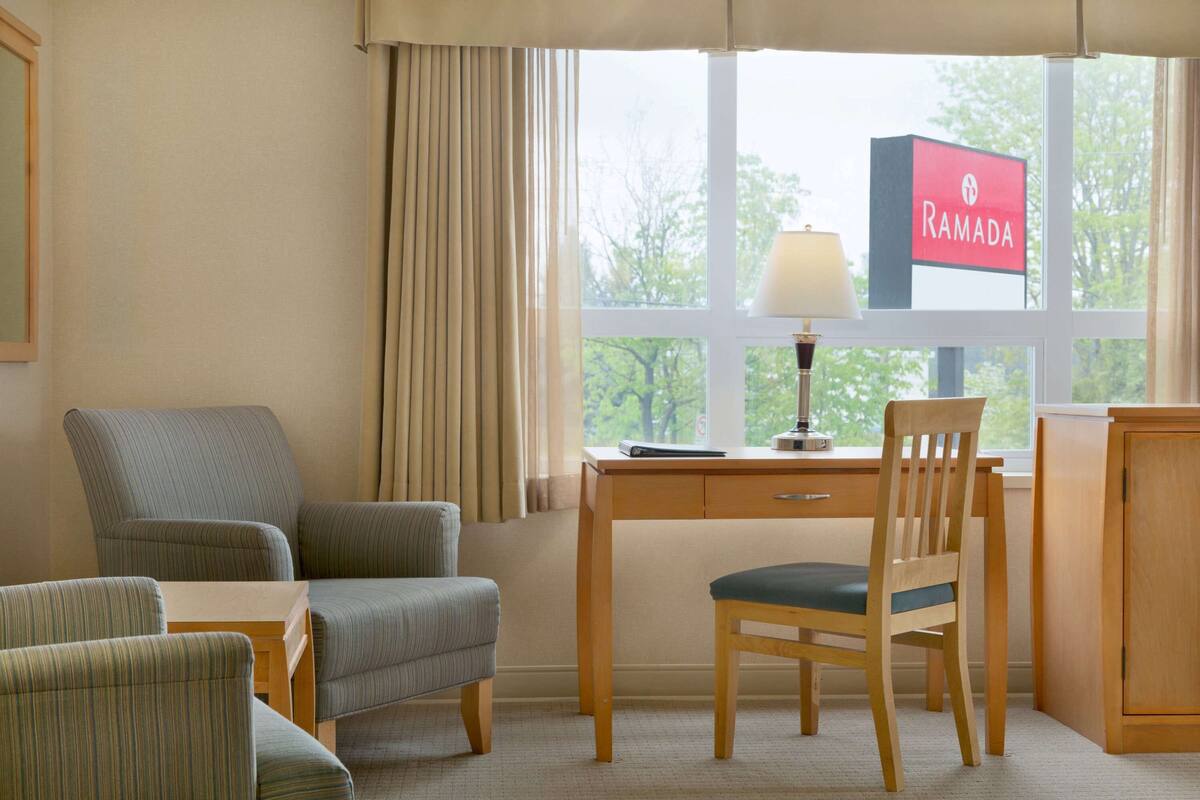 Habitación de hotel con escritorio, dos sillas, lámpara y vista a los árboles desde la ventana.