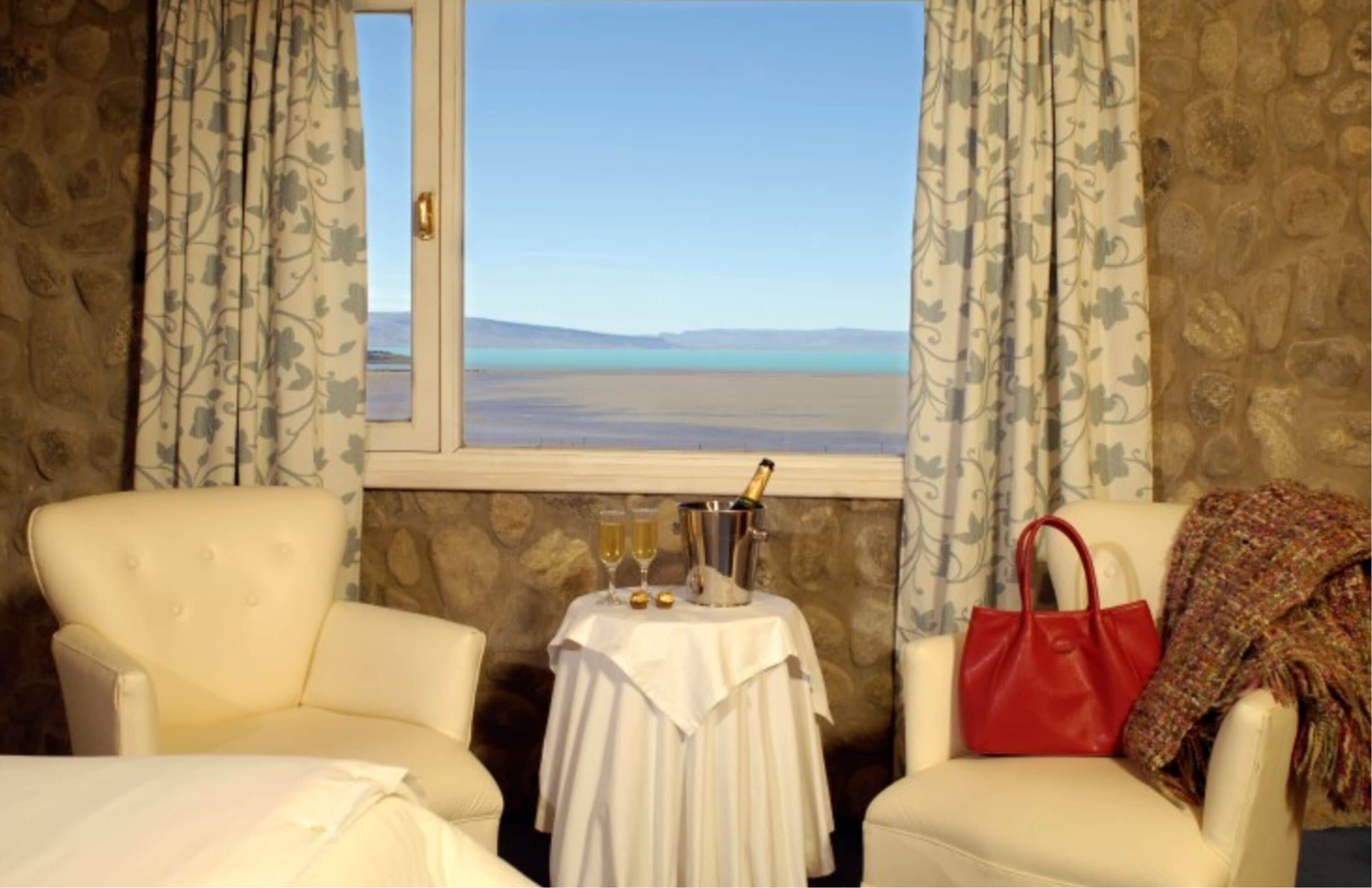 Una habitación con dos sillas, una mesa con una botella y vasos, y vista a un cuerpo de agua a través de una ventana.