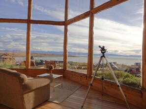 Eine Holzhütte mit einem großen Fenster mit Blick auf einen See und Berge. Im Inneren befindet sich eine Couch und ein Teleskop auf einem Stativ.