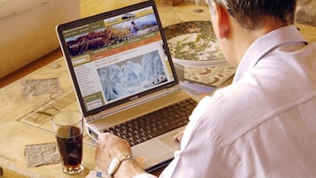 Une personne est assise à une table en bois et utilise un ordinateur portable. Sur la table, on aperçoit également un verre contenant une boisson sombre, un carnet et un stylo.